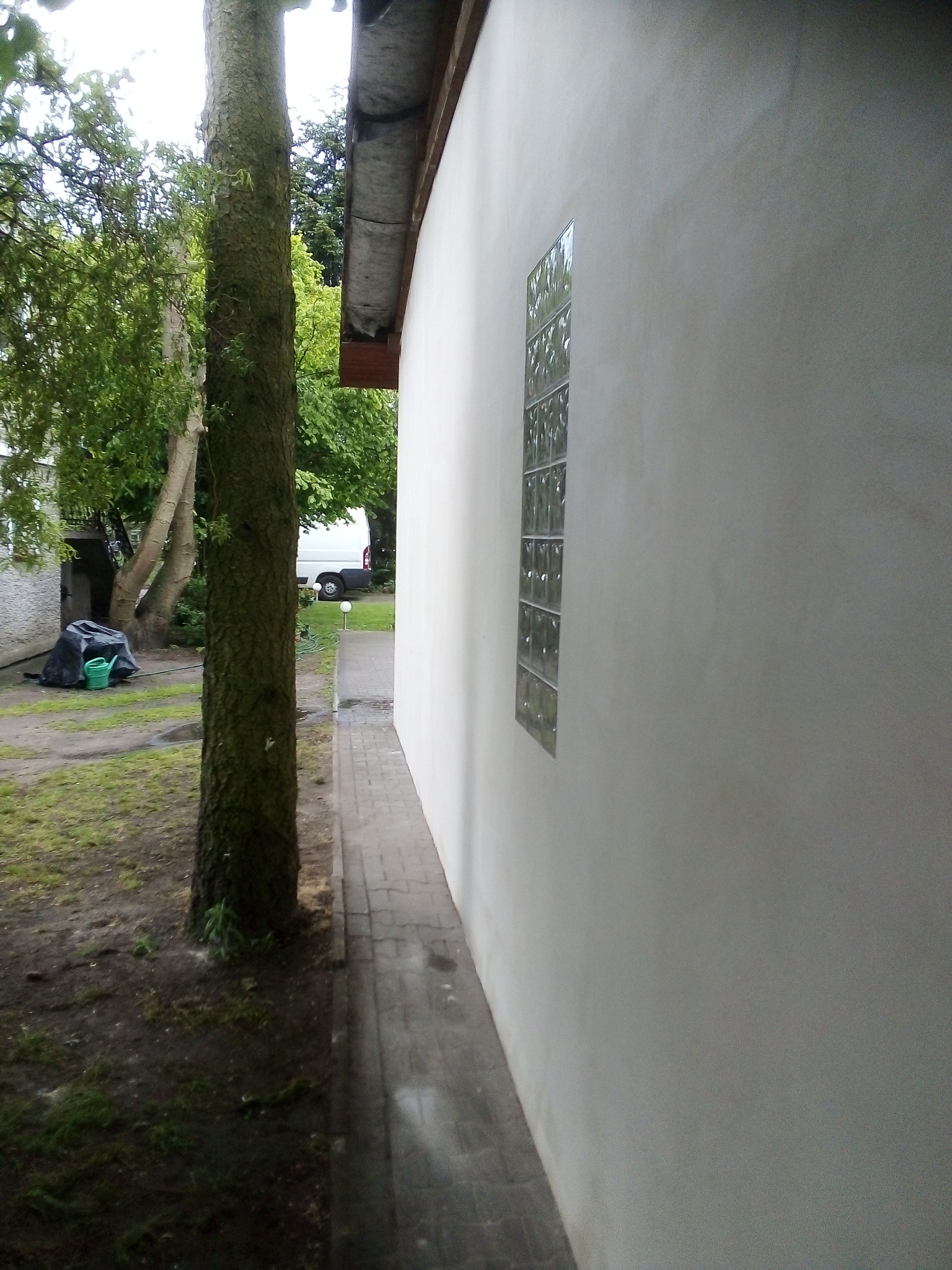 Świeżo otynkowana ściana zewnętrzna budynku z wstawionym oknem z luksferów, widoczna z perspektywy chodnika i pnia drzewa, z zarysem podwórka i zaparkowanego busa w tle.
