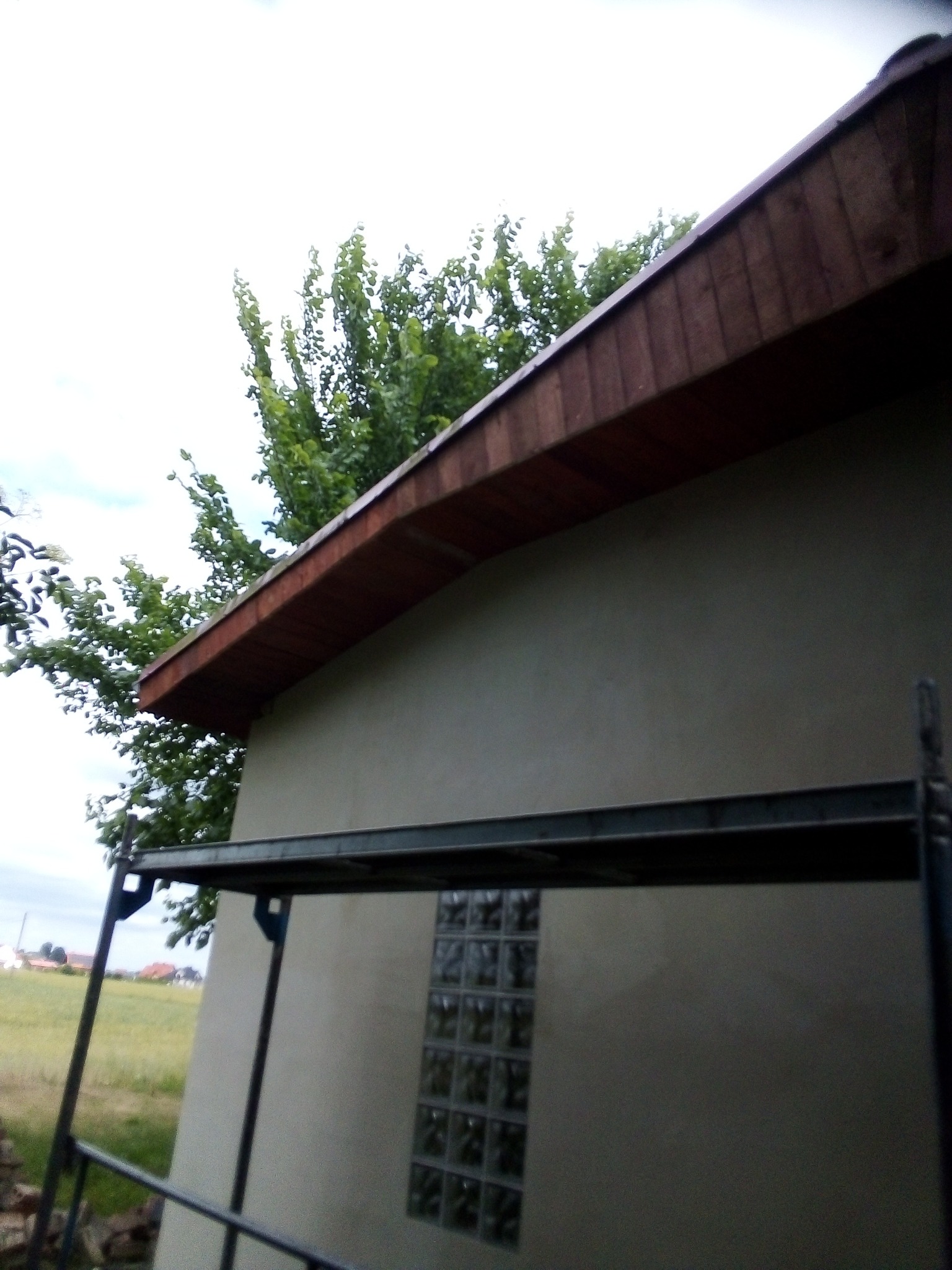 Elewacja budynku z oknem z luksferów, widoczna z poziomu rusztowania, z brązowym drewnianym okapem i zielonymi drzewami w tle.