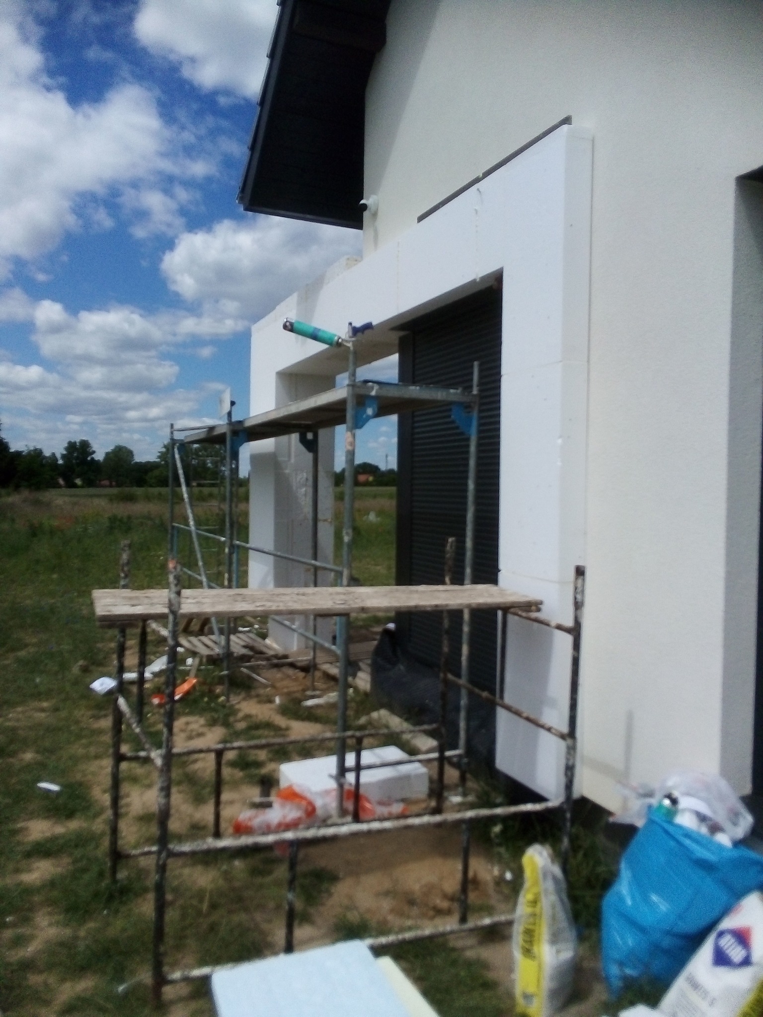 Budynek w trakcie ocieplania elewacji, widoczne rusztowanie, styropianowe obramowanie okna i worki z klejem do ociepleń.