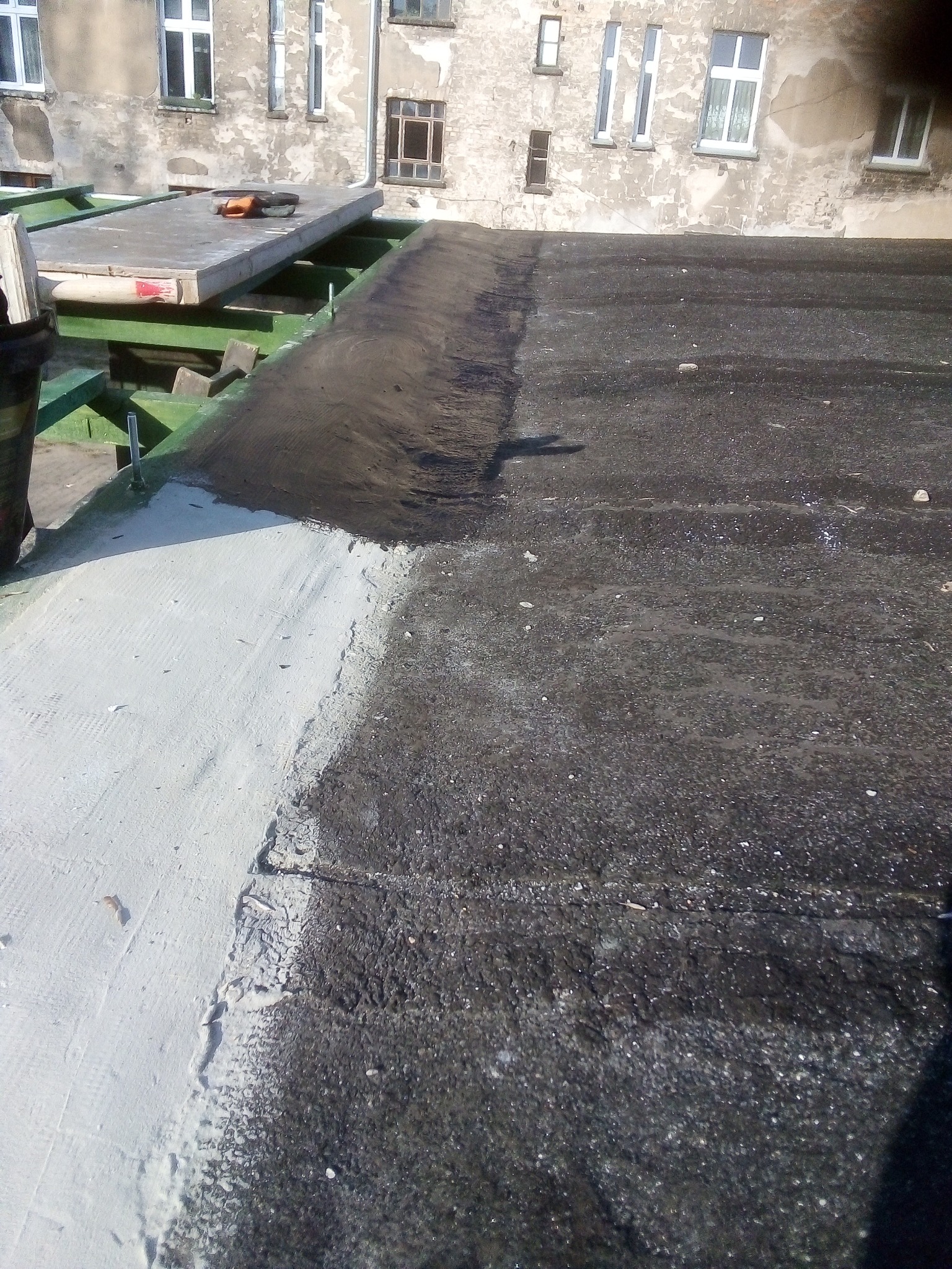 Fragment dachu z częściowo nałożoną białą warstwą izolacyjną, widoczna stara, czarna powierzchnia i fragment konstrukcji dachu z narzędziami w tle.