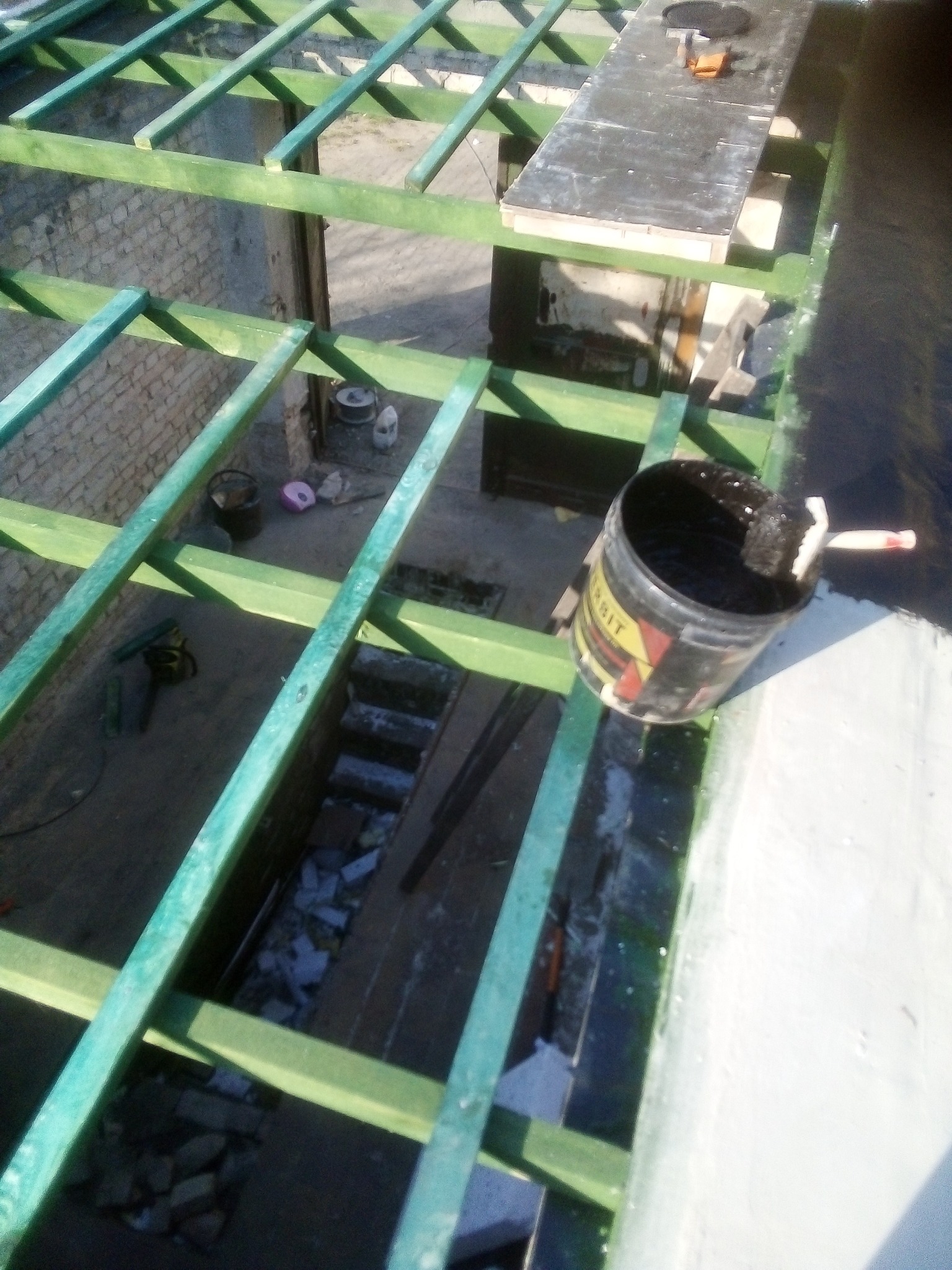 Widok z góry na konstrukcję dachu z drewnianych belek pomalowanych na zielono, z widocznym wiadrem z czarną substancją i pędzlem, oraz fragmentem białej powierzchni obok, sugerujący prace...