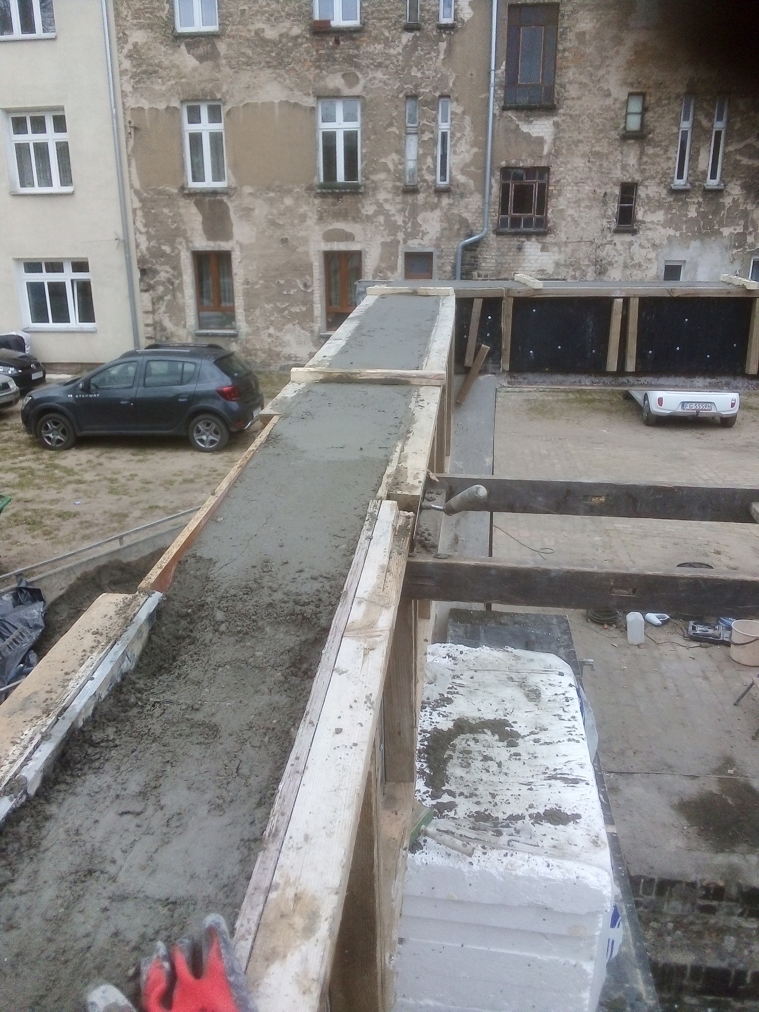 Wylewanie betonu w szalunku na elewacji budynku, widoczne drewniane konstrukcje i fragment czerwonej rękawicy roboczej.