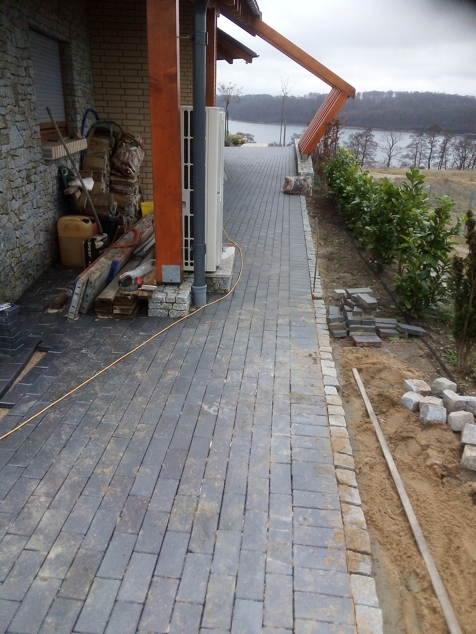 Częściowo ułożona kostka brukowa wzdłuż ścieżki prowadzącej w kierunku jeziora, z widocznymi stertami kostki i narzędziami budowlanymi, obok domu z kamienną elewacją.