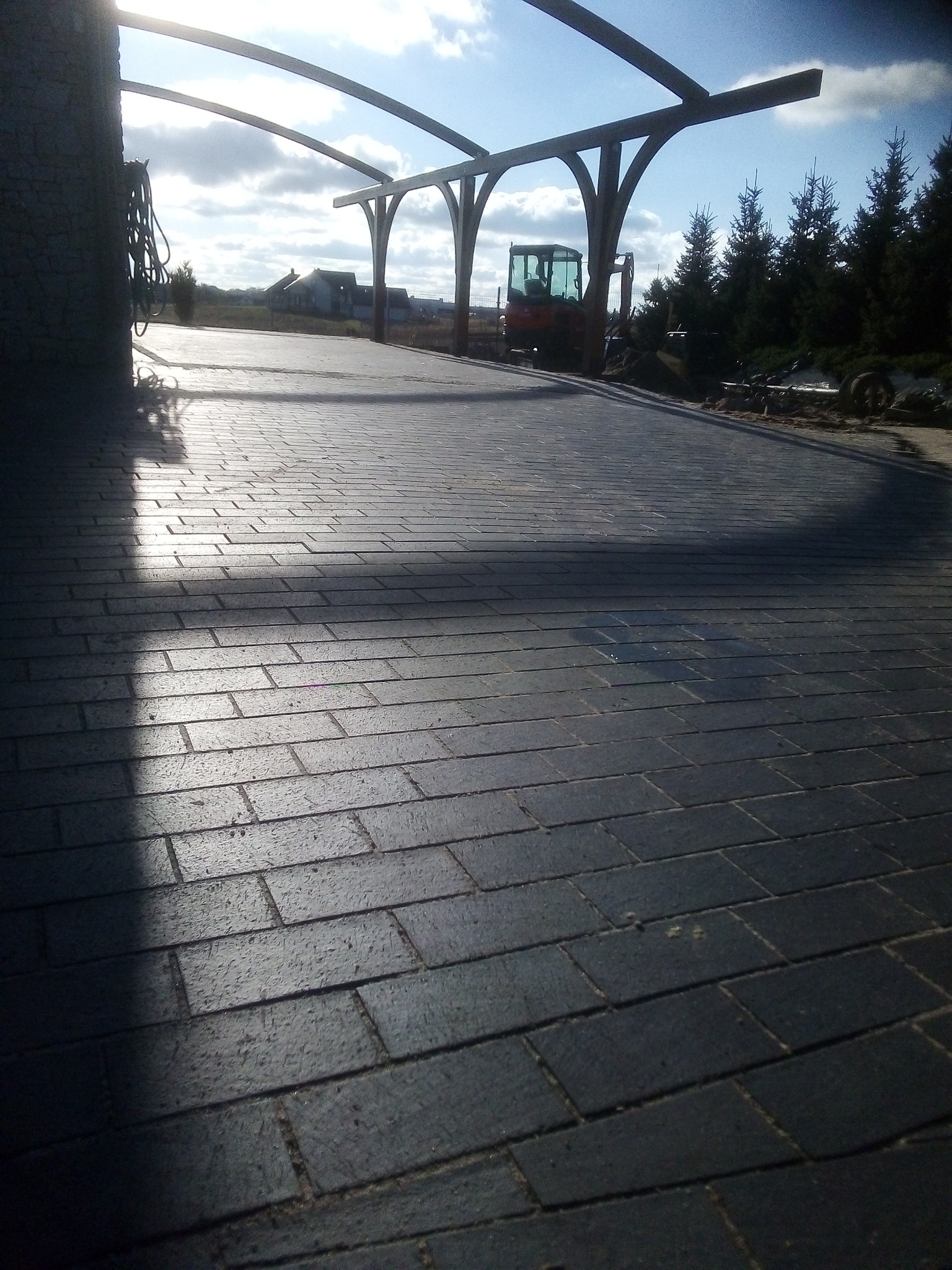 Ułożona, ciemnoszara kostka brukowa pod drewnianą konstrukcją z podcieniami, w tle widoczny mały traktor i drzewa iglaste.