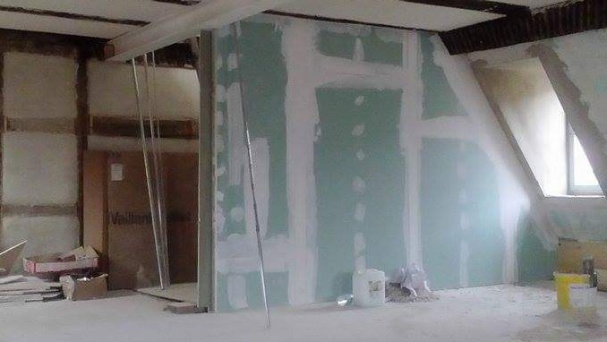 Wnętrze poddasza w trakcie prac wykończeniowych: ściany z płyt kartonowo-gipsowych z widocznymi łączeniami szpachlowanymi, metalowe profile, kartony i pojemniki z materiałami budowlanymi na podłodze.