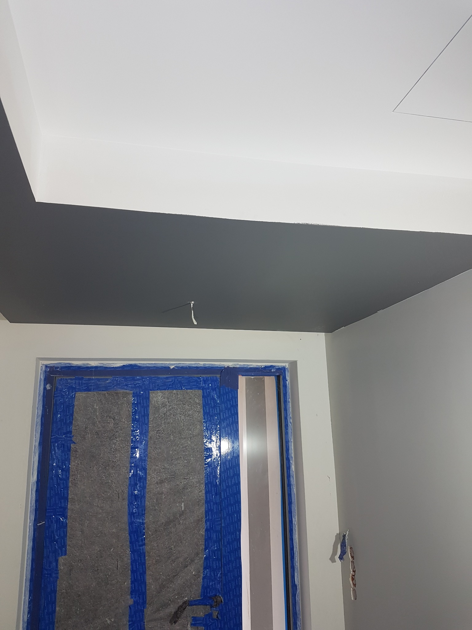 Ciemnoszary sufit pomalowany w nowym mieszkaniu, z zabezpieczonymi folią i taśmą malarską drzwiami w tle, widoczne niedociągnięcia przy krawędzi sufitu.
