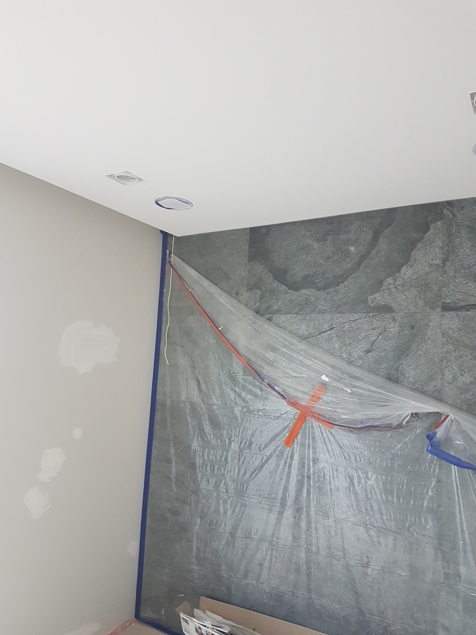 Przygotowanie do prac wykończeniowych w pomieszczeniu: ściana z szarych płytek zabezpieczona folią malarską z pomarańczową taśmą krzyżową, obok ściana zagruntowana z widocznymi plamami szpachli...