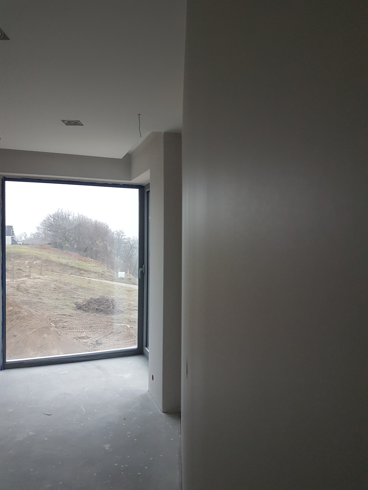 Wnętrze w trakcie prac wykończeniowych: świeżo pomalowane ściany w odcieniach szarości, widoczne surowe wykończenie i duże okno z widokiem na zewnątrz, zarys instalacji elektrycznej na suficie.