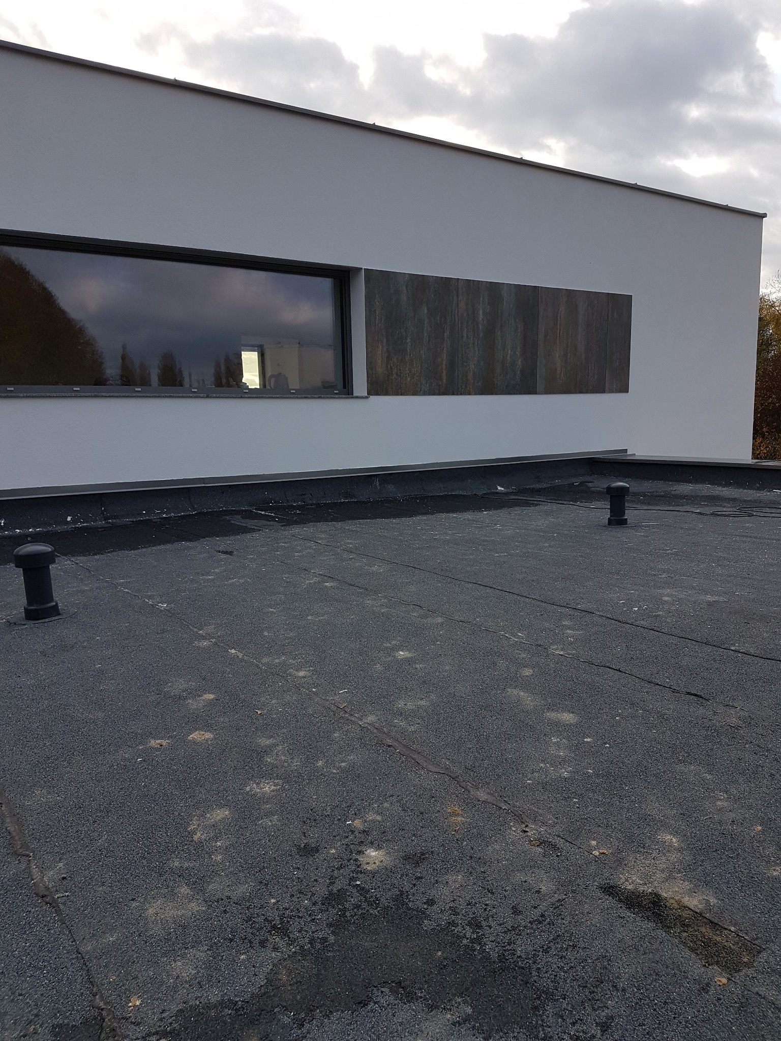 Widok z góry na płaski dach pokryty papą z dwoma odpowietrznikami, na tle ściany budynku z oknem i elementem dekoracyjnym z paneli imitujących drewno.