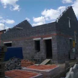 Surowy stan domu w budowie z szarych bloczków, widoczne okna i zarys dachu, w tle zabudowania sąsiedzkie, na pierwszym planie materiały budowlane.