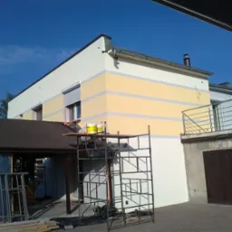 Dwukondygnacyjny dom z nową, dwukolorową elewacją w odcieniach bieli i żółci, z widocznymi rusztowaniami i pojemnikami z farbą na dachu wiaty garażowej. Świeżo nałożony tynk strukturalny.