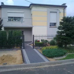 Dwukondygnacyjny dom z elewacją w odcieniach szarości i beżu, z szarą kostką brukową prowadzącą do furtki z numerem 2, obok widoczna pryzma piasku.