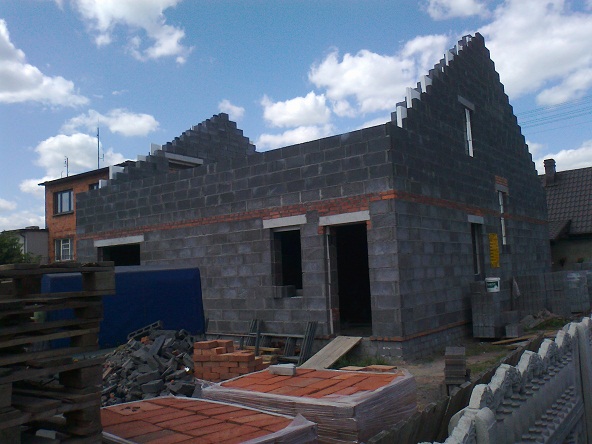 Surowy stan domu w budowie z szarych bloczków, widoczne okna i zarys dachu, w tle zabudowania sąsiedzkie, na pierwszym planie materiały budowlane.