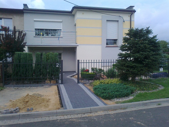 Dwukondygnacyjny dom z elewacją w odcieniach szarości i beżu, z szarą kostką brukową prowadzącą do furtki z numerem 2, obok widoczna pryzma piasku.