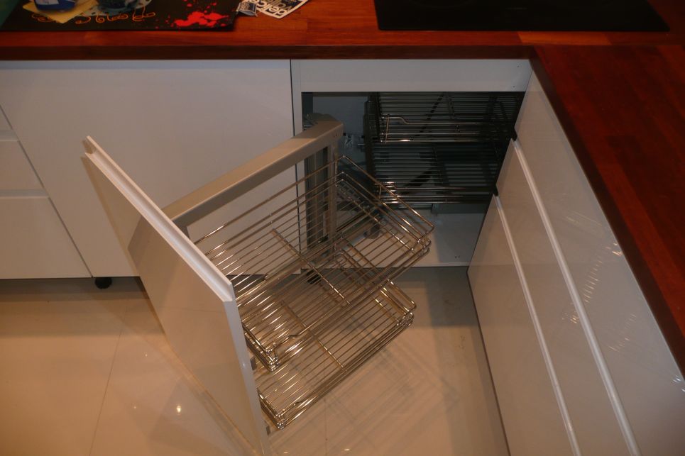 Otwarta szafka narożna z systemem wysuwnych, metalowych koszy w nowoczesnej kuchni z białymi frontami i drewnianym blatem.