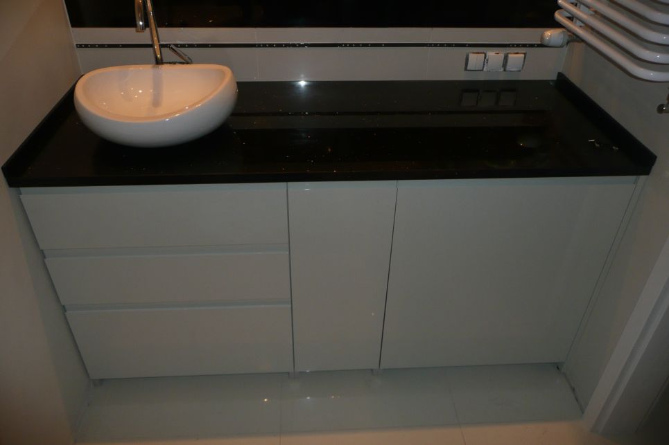 Nowoczesna umywalka nablatowa w kształcie misy, zamontowana na szafce z szufladami i drzwiczkami w kolorze jasnoszarym, z czarnym, błyszczącym blatem z delikatnym brokatem. Widoczne białe płytki...