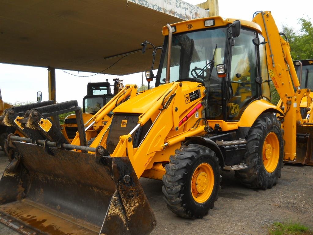 Żółta maszyna budowlana JCB 3CX z łyżką z przodu, stojąca na placu pod zadaszeniem. Widoczne inne maszyny w tle.