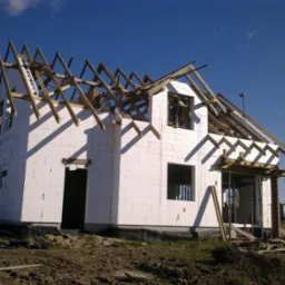 Niedokończony budynek jednorodzinny w systemie Thermomur z widoczną konstrukcją więźby dachowej i niezabudowanymi otworami okiennymi, z ziemią wok&oacute;ł budynku.