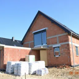 Surowy budynek z czerwonej cegły z zamontowanymi oknami i roletami zewnętrznymi, widoczne worki z materiałami budowlanymi, garaż w trakcie budowy, data na zdjęciu.