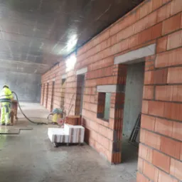 Wnętrze budynku w trakcie murowania ścian z czerwonej cegły dziurawki, widoczne otwory okienne z nadprożami, pracownik w odblaskowej odzieży używa urządzenia do obróbki betonu, w tle surowy...