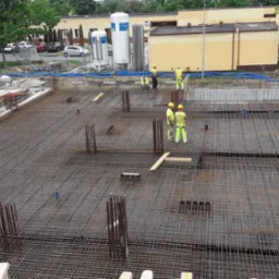 Siatka zbrojeniowa na placu budowy, widoczni pracownicy w odblaskowych ubraniach, przygotowania do wylewania fundamentów, w tle budynki i drzewa.