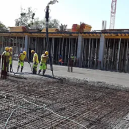 Wylewanie betonu na siatkę zbrojeniową na budowie budynku żelbetowego; pracownicy w żółtych ubraniach rozprowadzają beton, w tle widoczne słupy konstrukcyjne i dźwig.