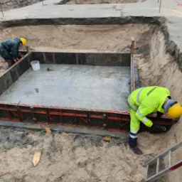 Dwóch robotników w żółtych kaskach i odblaskowych ubraniach montuje szalunek z czerwonymi elementami do wylania fundamentu w wykopie zabezpieczonym taśmą ostrzegawczą, w tle hałdy piasku i sprzęt...