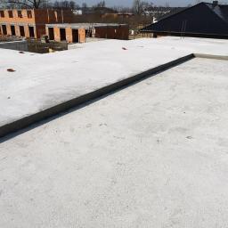 KBD KOMPLEKSOWA BUDOWA DOMÓW JAROSŁAW KOCLĘGA - Zalany betonowy strop na budowie domu jednorodzinnego, w tle budynek w trakcie wznoszenia z czerwonej cegły, widoczne stalowe pręty zbrojeniowe.