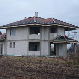 KBD KOMPLEKSOWA BUDOWA DOMÓW JAROSŁAW KOCLĘGA - Dwukondygnacyjny dom w trakcie budowy z pustaków, z częściowo ukończonym dachem z czerwonej dachówki, widoczne rusztowania i pracownik na dachu, szare niebo w tle.