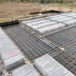 Zbrojony fundament z betonowych bloczków, widok z góry na siatkę stalową i bloczki z różowymi oznaczeniami, w tle wykop ziemny i drzewa.