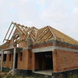 Wznoszenie ścian z czerwonej cegły i konstrukcji dachu z drewna na budowie domu jednorodzinnego w Prochowicach, widoczne zbrojenia nad otworami okiennymi.