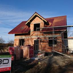 KBD KOMPLEKSOWA BUDOWA DOMÓW JAROSŁAW KOCLĘGA - Wznoszony dwukondygnacyjny budynek z czerwonej cegły z częściowo ułożonym dachem z czerwonej dachówki, widoczne rusztowanie, palety cegieł Wienerberger oraz baner firmy budowlanej KBO.