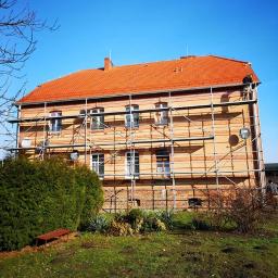 KBD KOMPLEKSOWA BUDOWA DOMÓW JAROSŁAW KOCLĘGA - Dwupiętrowy budynek z cegły i tynkiem w trakcie renowacji, otoczony rusztowaniem, z widocznym pracownikiem na dachu i zielenią w otoczeniu.