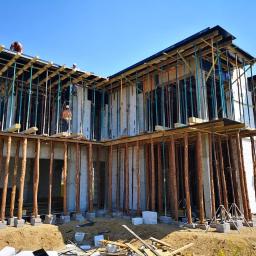 KBD KOMPLEKSOWA BUDOWA DOMÓW JAROSŁAW KOCLĘGA - Surowy stan budowy dwukondygnacyjnego domu z widocznymi betonowymi ścianami, drewnianymi stemplami i metalowymi podporami, robotnicy na dachu, błękitne niebo w tle.