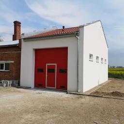 KBD KOMPLEKSOWA BUDOWA DOMÓW JAROSŁAW KOCLĘGA - Nowo wybudowany, biały budynek z czerwoną bramą garażową segmentową, połączony z ceglanym budynkiem z kominem, widok z zewnątrz na tle pola.