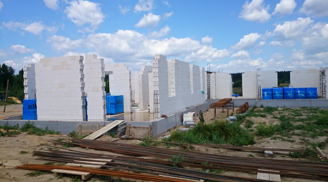 Wznoszenie ścian domu z białych bloczków silikatowych na fundamencie. Na pierwszym planie stalowe pręty zbrojeniowe. W tle błękitne niebo z obłokami.