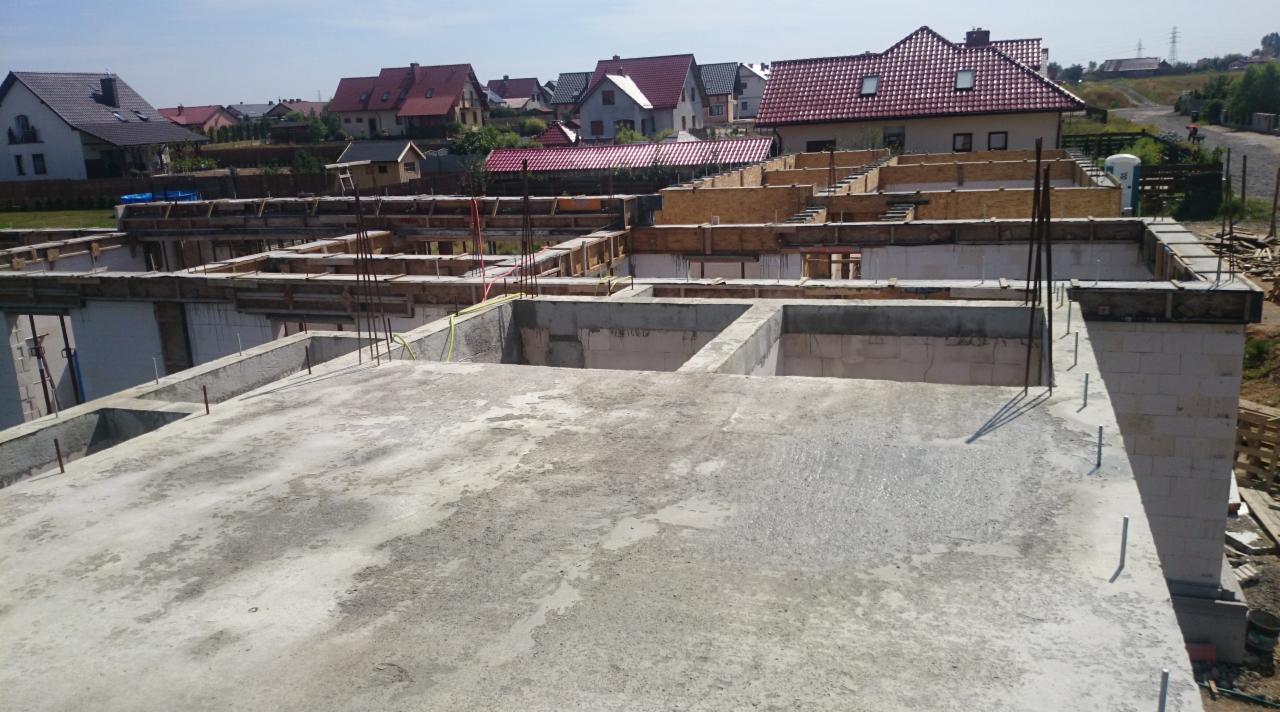 Wznoszenie ścian fundamentowych z bloczków i szalunków, widok z góry na plac budowy z domami w tle, słoneczny dzień.