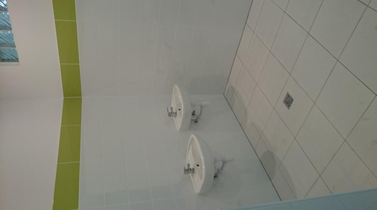 Dwie białe umywalki zamontowane na białej, wykafelkowanej ścianie w łazience. Widoczny zielony pas płytek i okno z luksferów.