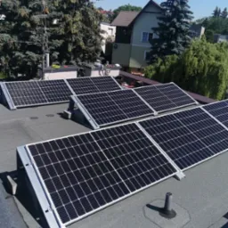 Panele słoneczne na płaskim dachu z widocznymi narzędziami i przewodami w tle, otoczone zielenią i budynkami mieszkalnymi w słoneczny dzień.