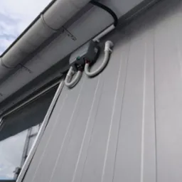 Szara skrzynka sterownicza z przewodami w peszlach przymocowana do metalowej ściany budynku, widok z dołu.