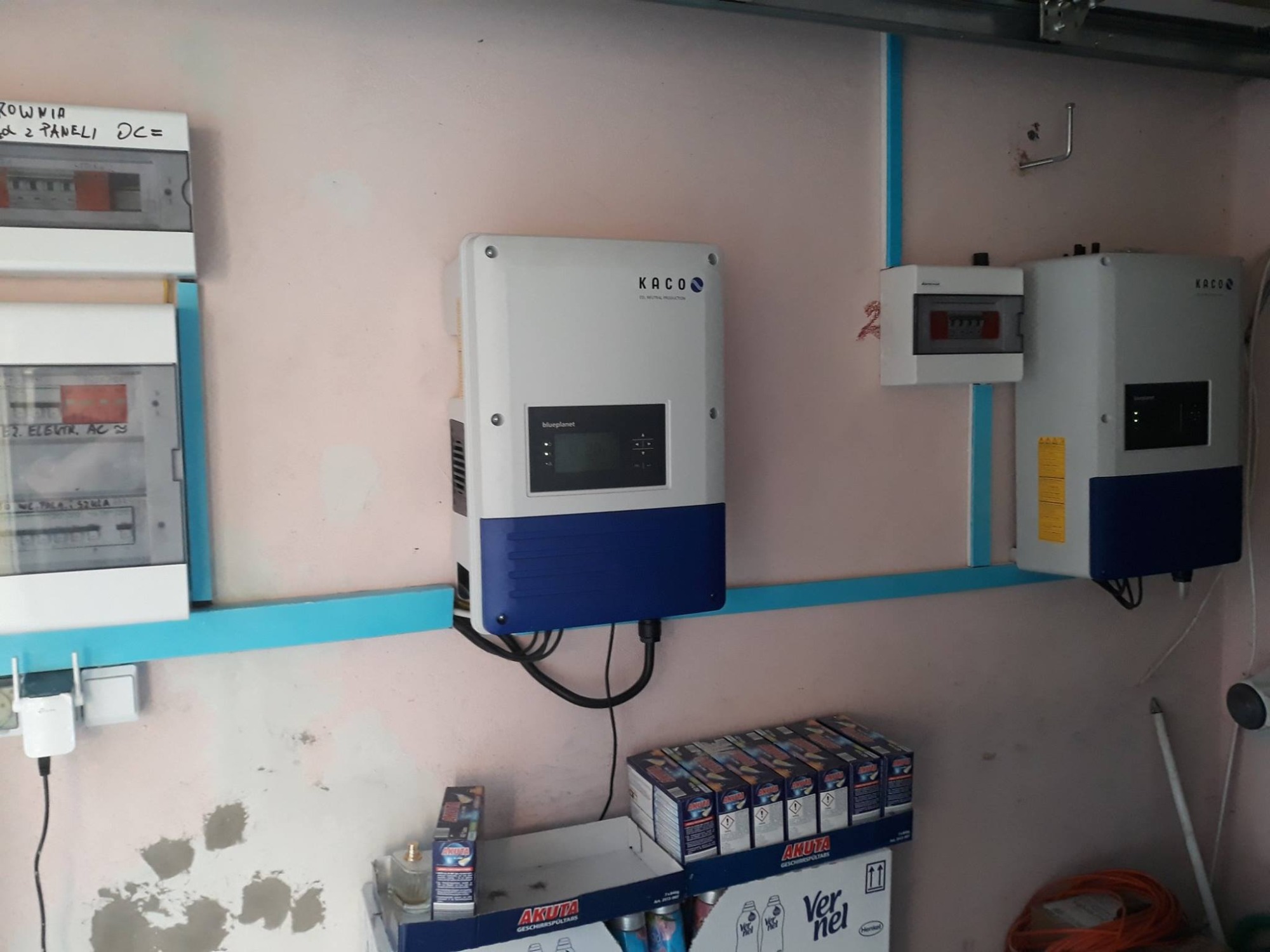Dwa inwertery fotowoltaiczne marki KACO zamontowane na ścianie w pomieszczeniu gospodarczym, obok skrzynki elektrycznej i półki z detergentami Akuta i płynem do płukania Ver nel.