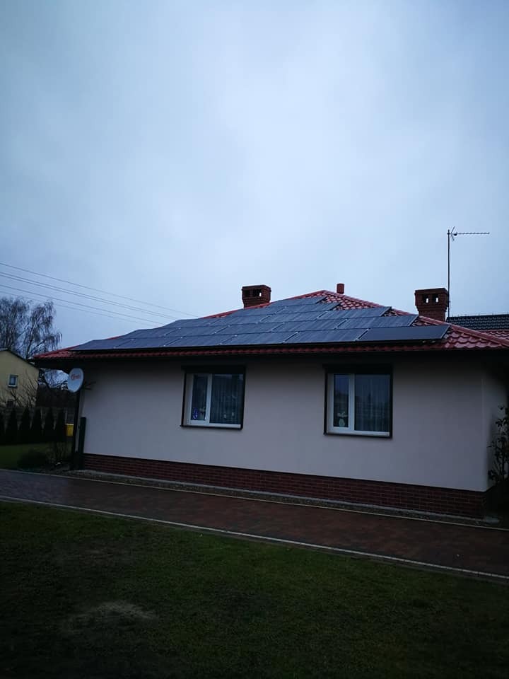 Dom jednorodzinny z panelami słonecznymi na dachu pokrytym czerwoną dachówką, widoczne kominy i antena satelitarna, trawnik przed budynkiem.