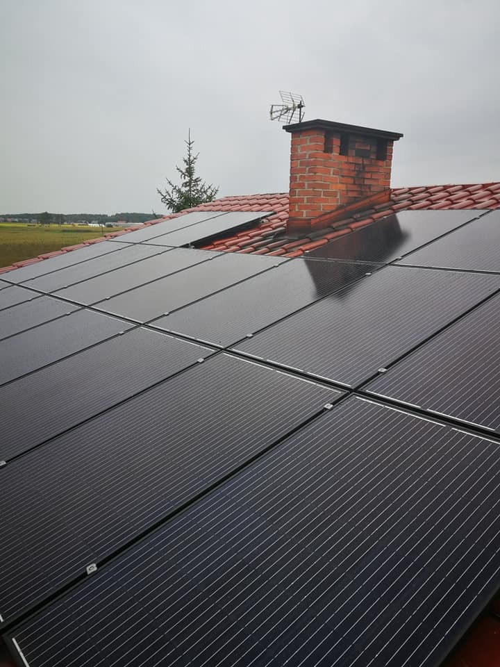 Panele słoneczne na dachu domu z ceglanym kominem i anteną telewizyjną w tle pochmurnego nieba i zielonego pola.