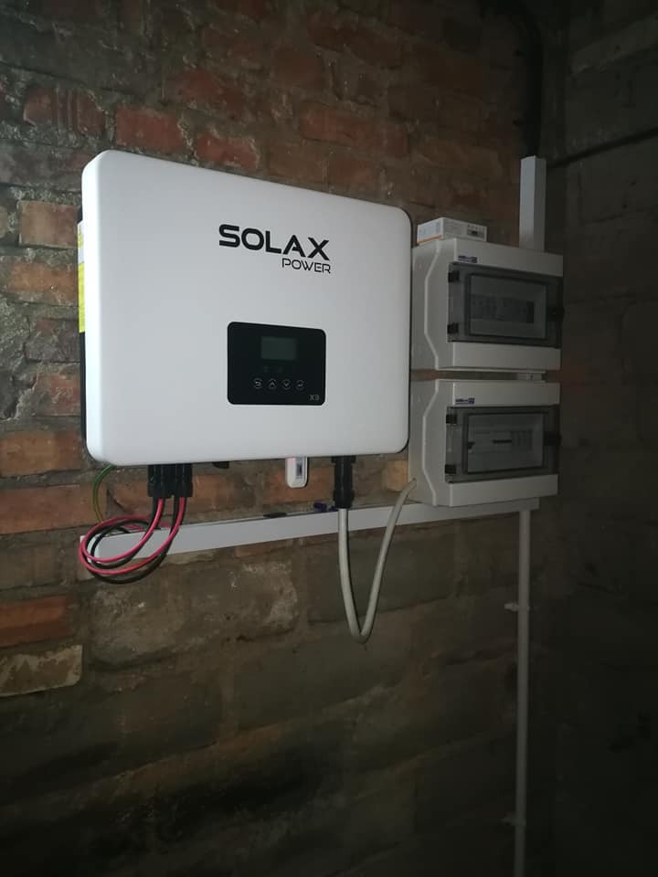 Biały inwerter Solax Power X3 zamontowany na ceglanej ścianie, z widocznymi przewodami i dwoma rozdzielnicami elektrycznymi po prawej stronie.