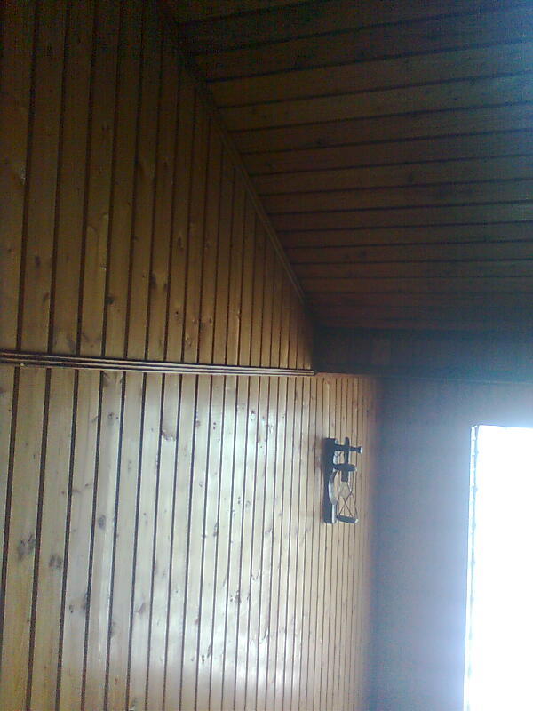 Drewniana boazeria ścienna i sufitowa z widocznymi rurami instalacji grzewczej oraz dekoracyjnym, metalowym elementem na ścianie.