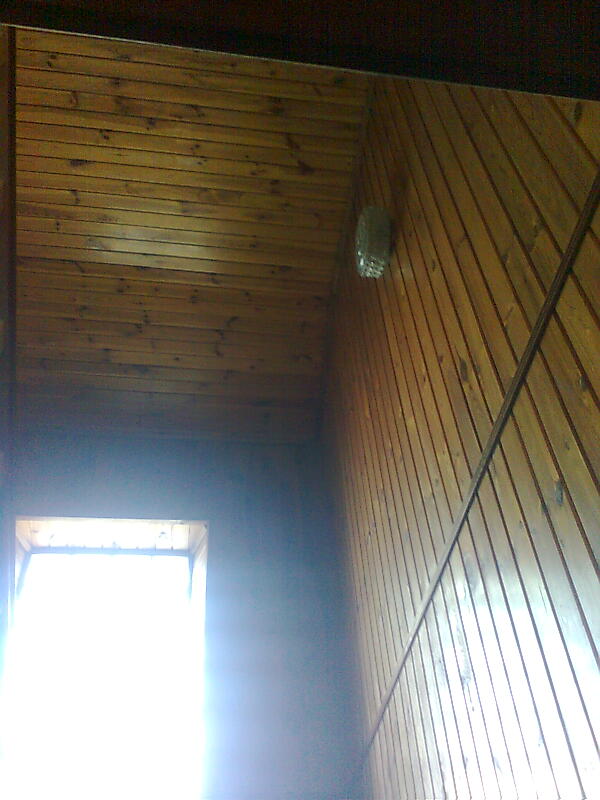 Wnętrze wykończone boazerią drewnianą, widok na ścianę i sufit pod kątem, z oknem w tle po lewej stronie, na ścianie widoczny element instalacji.