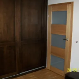 Jasne, drewniane drzwi wewnętrzne z przeszkleniami, zamontowane w pomieszczeniu obok ciemnej szafy przesuwnej. Widoczne wykończenie podłogi.