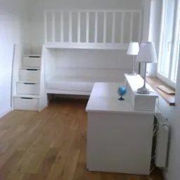 Białe łóżko piętrowe z biurkiem i schodami z szufladami w pokoju z drewnianą podłogą i oknem, oświetlone dwiema lampkami.