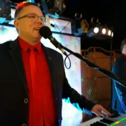 Mężczyzna w czarnej marynarce i czerwonej koszuli śpiewa do mikrofonu, grając na keyboardzie podczas występu na scenie z oświetleniem.