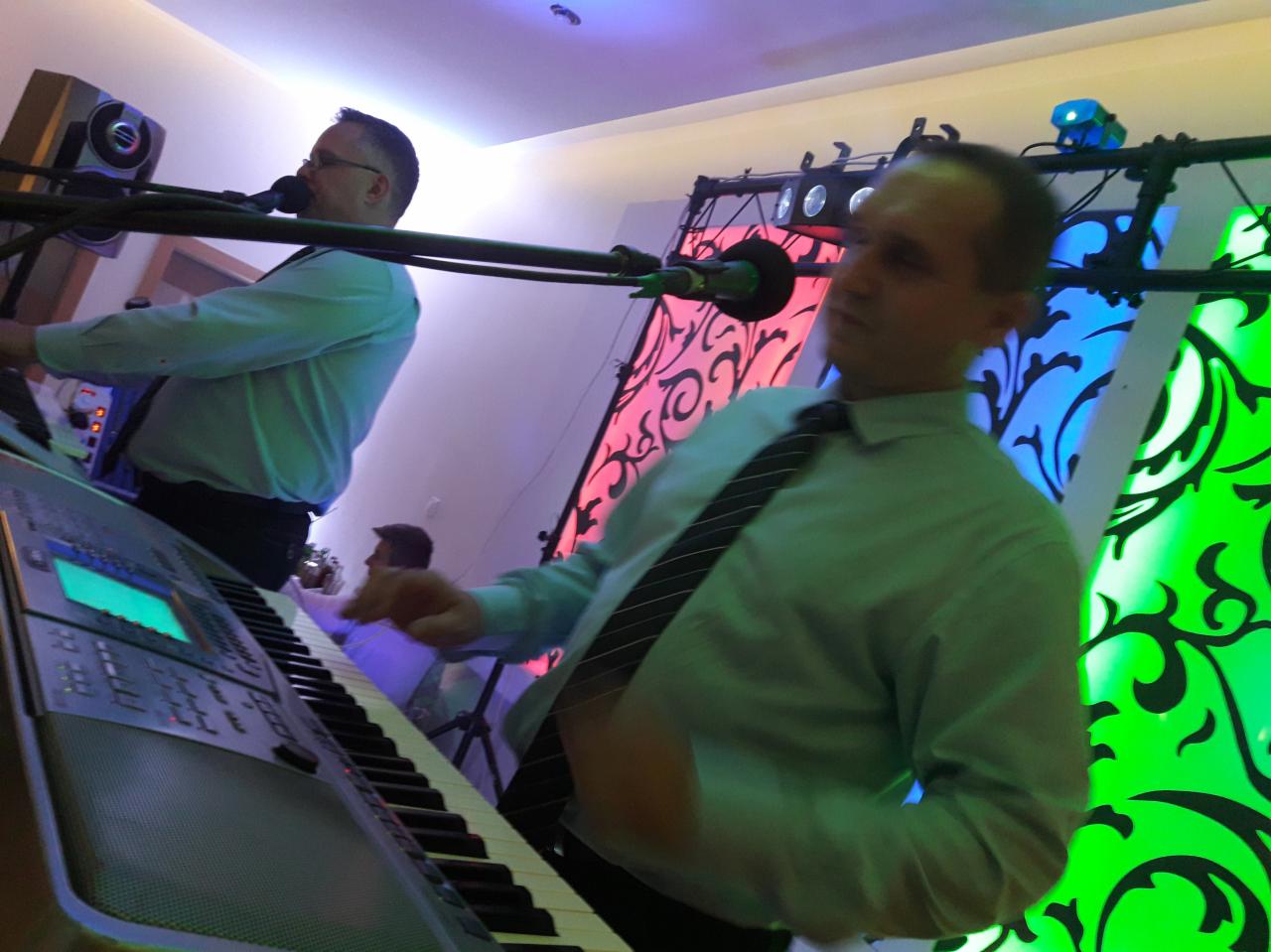 Dwóch mężczyzn grających na keyboardach i śpiewających do mikrofonów podczas występu, z dekoracyjnym tłem oświetlonym na czerwono, niebiesko i zielono.